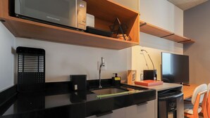A modern kitchen with a black countertop, a stainless steel microwave, a sink, and a dishwasher. There is a wooden shelf above the microwave and a television in the background. - Flat Moderno Perto do Metr e do Parque Ibirapuera (São Paulo)