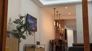 A modern living room with a flat-screen TV mounted on the wall, a dining area with a wooden table and chairs, and a large plant in the corner.
