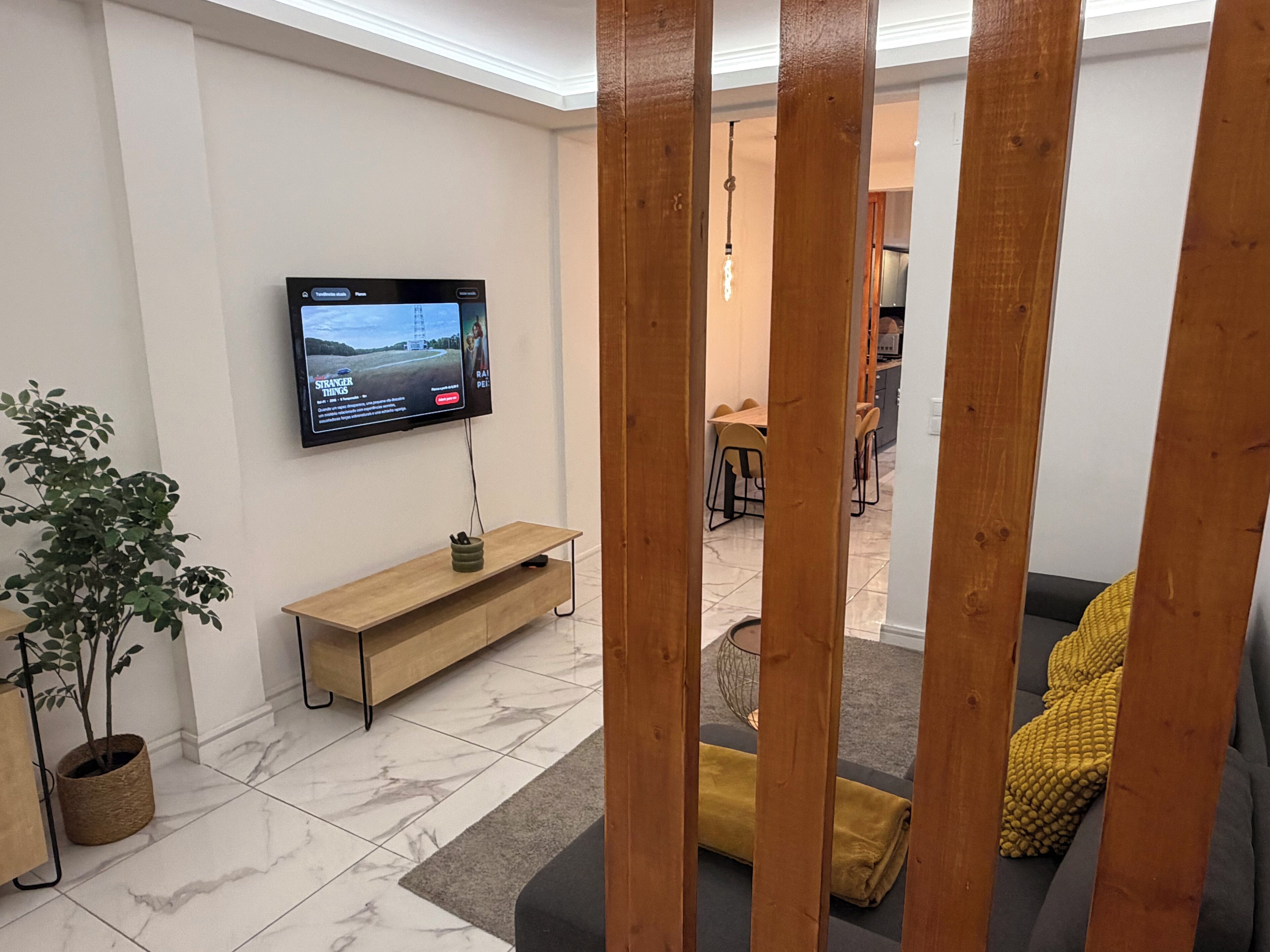 A living room with a TV mounted on the wall, a wooden bench, a plant, and a sofa with a yellow throw.