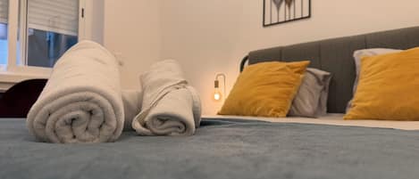 A neatly made bed with two rolled towels, a gray headboard, and a mustard-colored pillow.