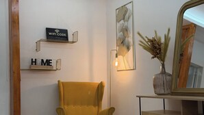 A room with a mustard yellow armchair, a small ottoman, a wooden cabinet, a mirror, a wall-mounted light, and a decorative wall piece.