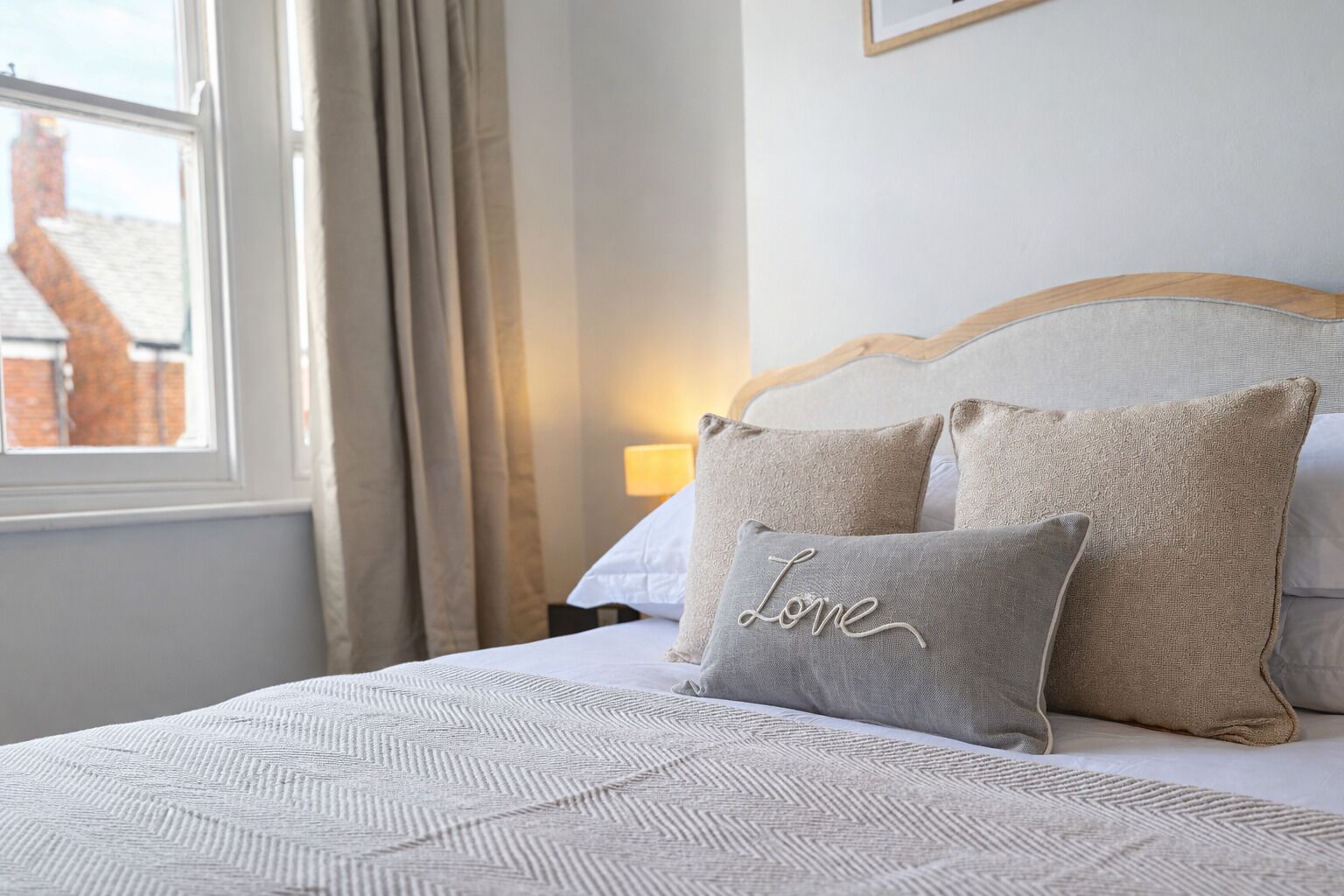 A neatly made bed with a patterned bedspread and two pillows, one with the word "Love" on it, in a room with a window and curtains.