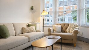Ein modernes Wohnzimmer mit Sofa, Sessel und Couchtisch, einer Stehlampe und Blick auf ein Backsteingebäude durch große Fenster.