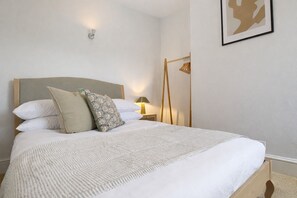 A neatly made bed with a grey headboard, white bedding, and patterned pillows. There is a bedside table with a lamp and a wooden hanger rack against the wall.