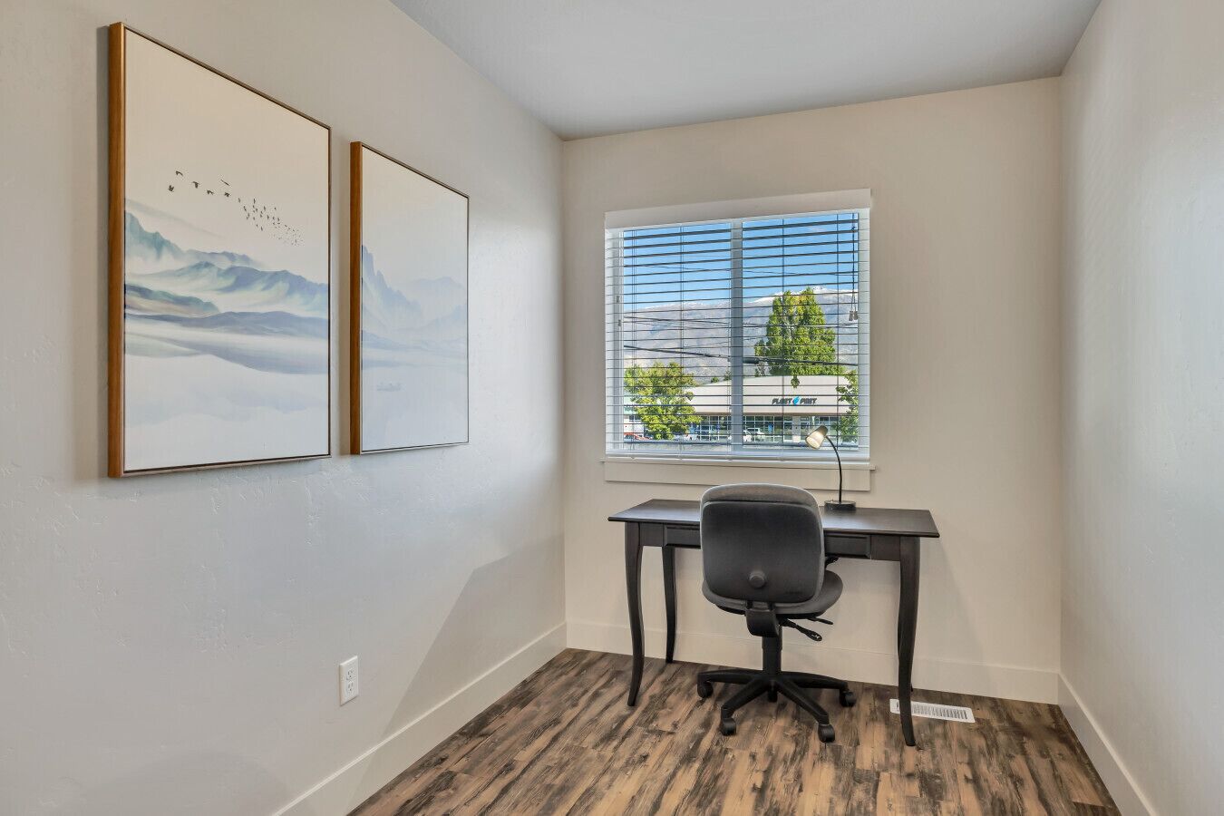 Un petit bureau comprenant un bureau et une chaise, deux œuvres d’art encadrées accrochées au mur et une fenêtre avec des stores.