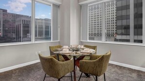 Un coin repas avec une table en verre pour quatre personnes, de grandes fenêtres offrant une vue sur la ville et un sol recouvert de moquette.