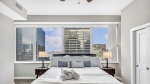 Quarto de hotel com uma cama grande, duas mesas de cabeceira com luminárias, vista de edifícios da cidade através de janelas amplas e uma porta à direita.