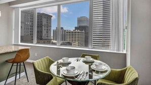 Uma sala de jantar moderna com uma mesa de vidro redonda, duas cadeiras estofadas em verde, e vista para edifícios da cidade através de grandes janelas.