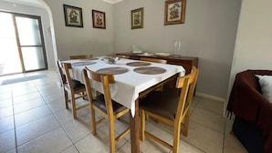 Un coin repas avec une table dressée pour quatre personnes, des chaises en bois et un banc. Des tableaux encadrés ornent le mur, et une porte donne sur l’extérieur.