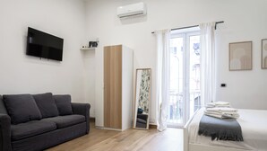 A modern room with a grey sofa, a bed with a white and grey blanket, a wooden wardrobe, a TV mounted on the wall, and a window with curtains. - CasaRÉ - One Bedroom Guest House, Sleeps 2 (Naples)