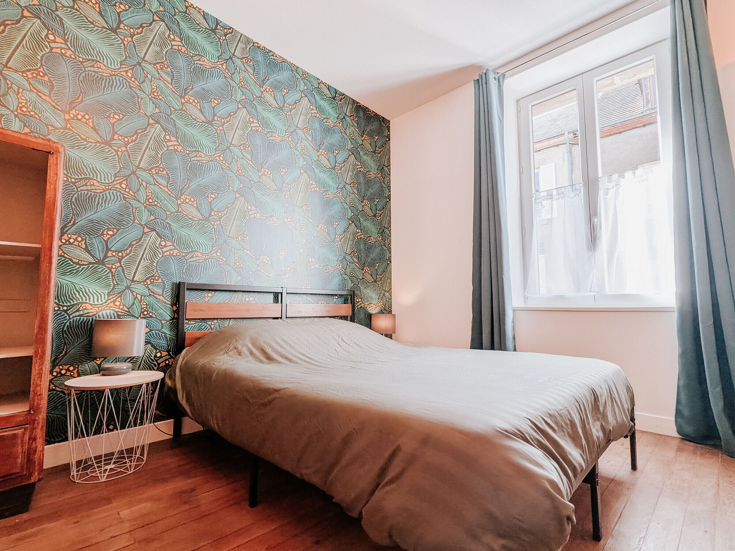 A bedroom with a patterned wall, a bed, a bedside table with a lamp, and a window with curtains.