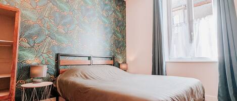 Un dormitorio con pared estampada, una cama, una mesita de noche con lámpara y una ventana con cortinas.