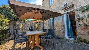 An outdoor patio with a table and chairs under a large umbrella, a stone wall with pineapple decorations, and a building entrance with a blue door. - Spacious cottage near Éguzon Lake (Orsennes)