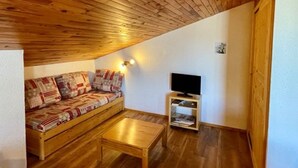 Una habitación acogedora con techo de madera, un sofá con cojines estampados, una mesita de madera y un televisor sobre una estantería.