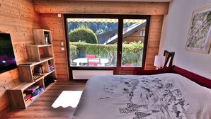 A bedroom with a large bed, wooden shelving, a TV, and a view of a green landscape through a sliding door.