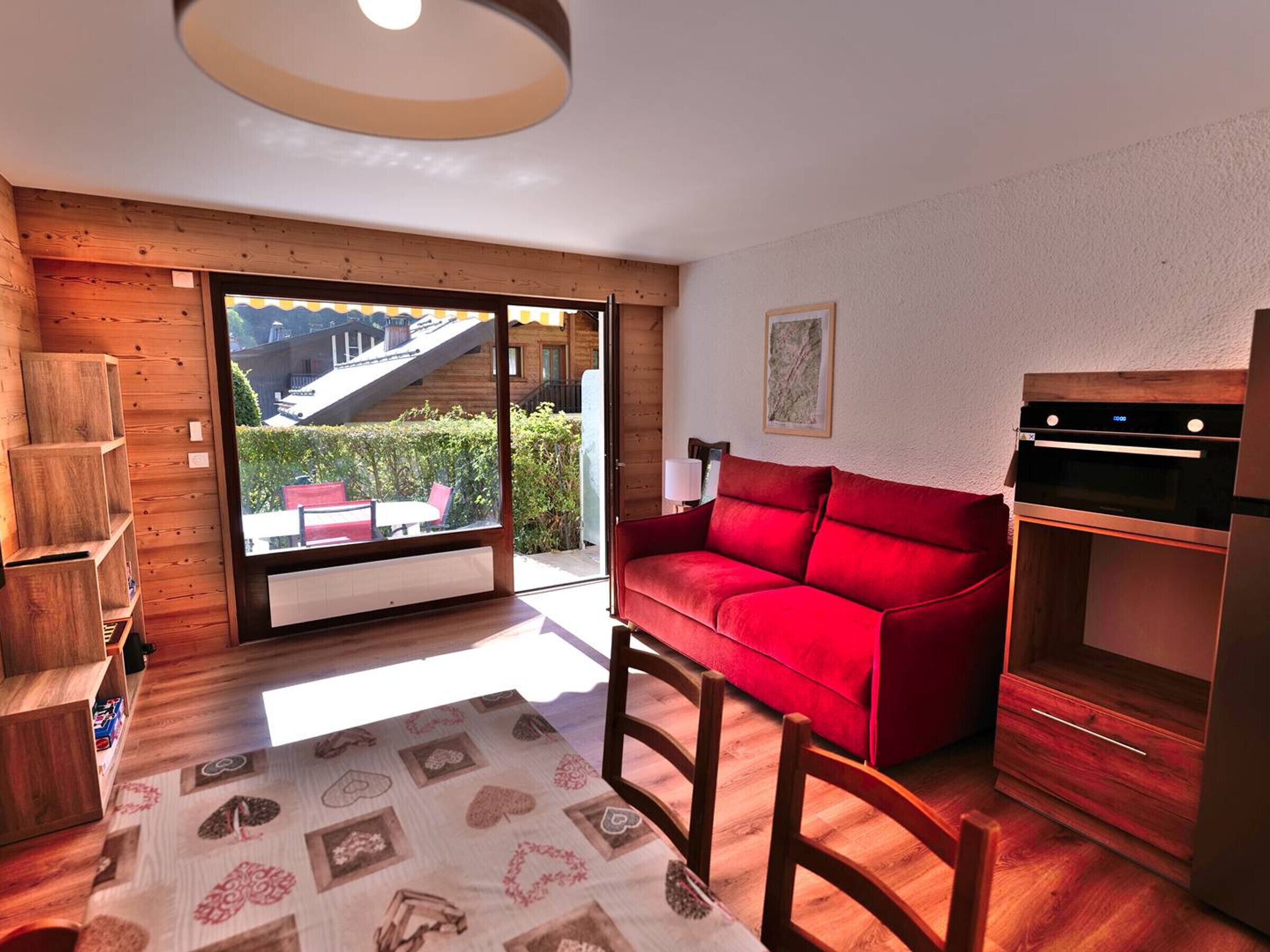 A cozy room with a red sofa, wooden shelves, a dining table with a patterned tablecloth, and a view of a building through a glass door.