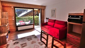 A cozy room with a red sofa, wooden shelves, a dining table with a patterned tablecloth, and a view of a building through a glass door.