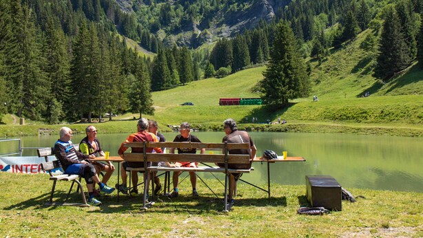 A group of people seated at a wooden table by a lake, with a mountainous backdrop.