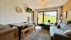 A cozy living room with a wooden table, bench, and sofa, a television, and a large window with a view of greenery.