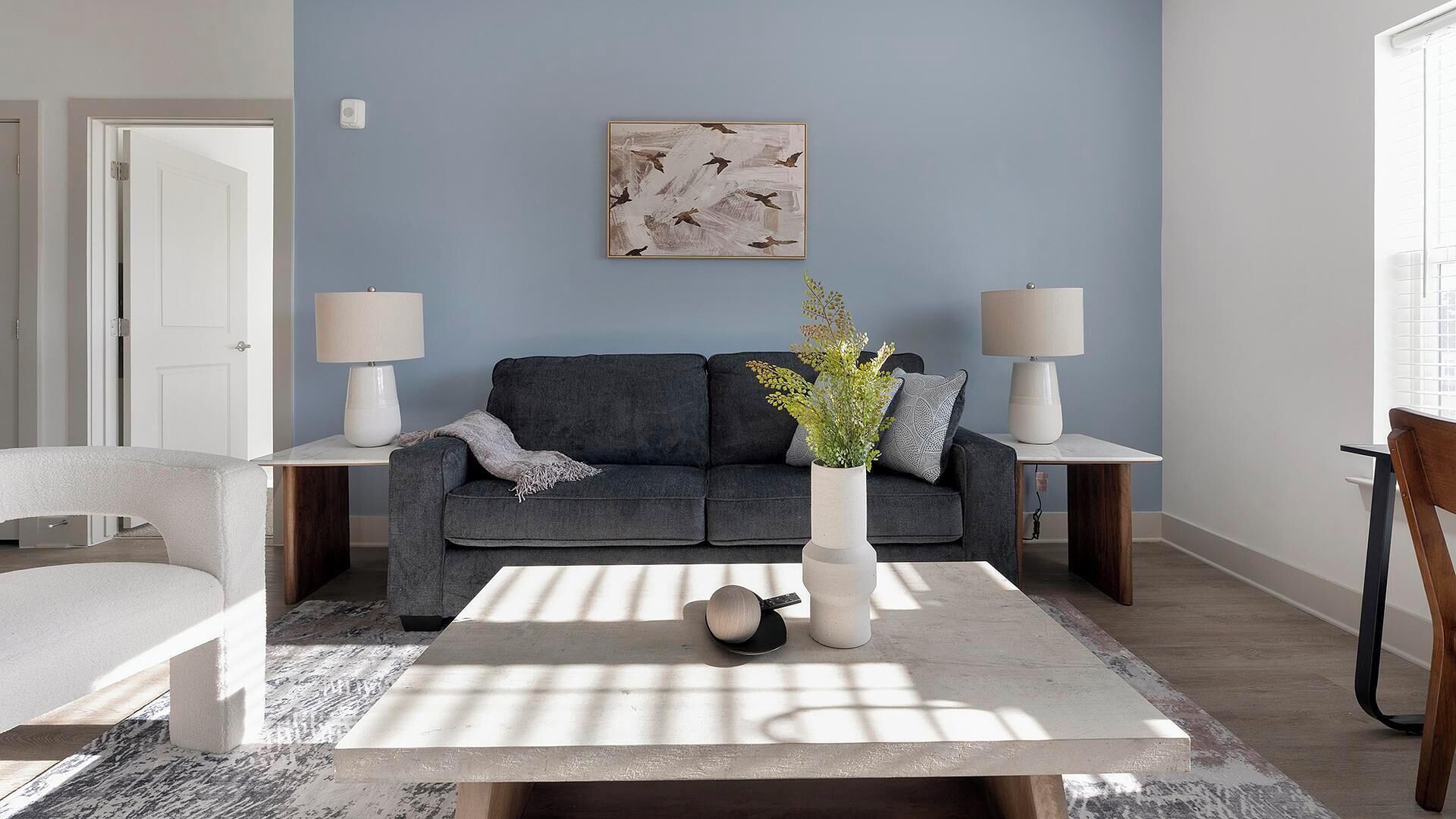 A living room with a dark grey sofa, two side tables with lamps, a coffee table with a vase and a plant, and a painting on the wall.