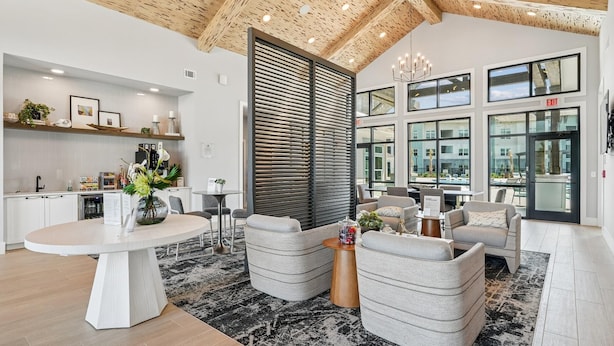 A modern hotel lobby with a high ceiling, a central seating area with sofas and a round table, a kitchen area with a counter and shelves, and large windows offering views of the outside. - Landing | Stunning 1BD, Gym, Yoga Studio (Myrtle Beach)