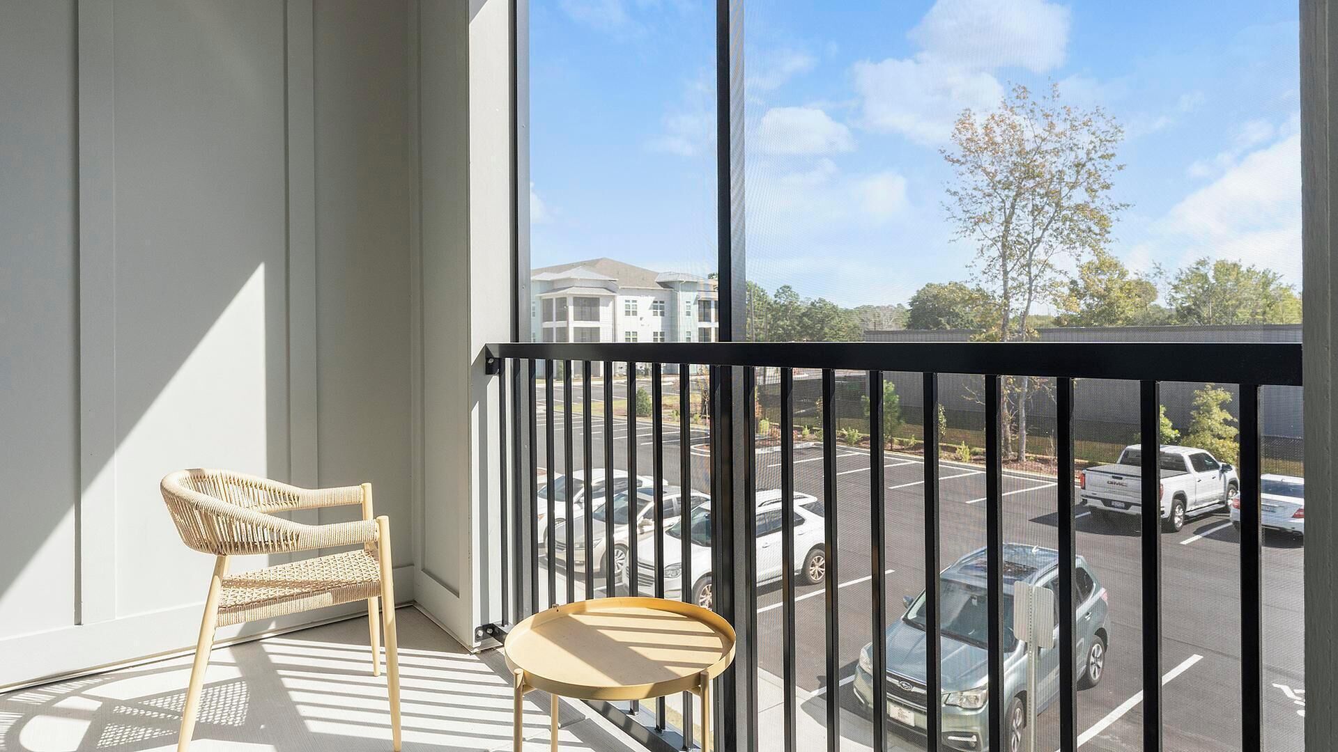 A balcony with a wicker chair and a small table, overlooking a parking lot and buildings.