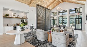A modern hotel lobby with a high ceiling, a central seating area with sofas and a round table, a kitchen area with a counter and shelves, and large windows offering views of the outside. - Landing | Stunning 1BD, Clubhouse, Yoga Studio (Myrtle Beach)