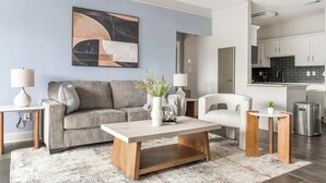 Ein modernes Wohnzimmer mit einem grauen Sofa, einem Couchtisch und einem weißen Sessel. An der Wand hängt ein Gemälde und im Hintergrund ist eine Küchenzeile zu sehen.