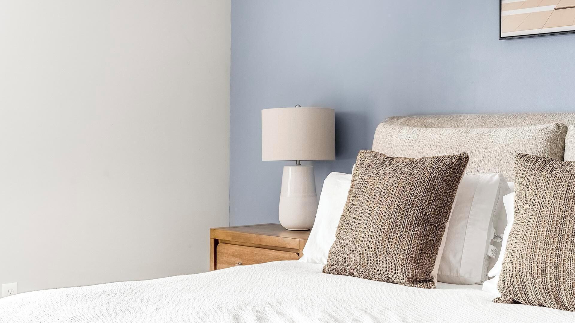 A neatly made bed with white bedding, a wooden nightstand with a lamp, and textured pillows.