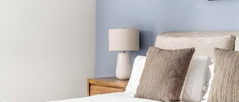 A neatly made bed with white bedding, a wooden nightstand with a lamp, and textured pillows.