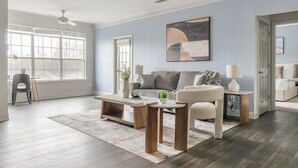 Ein geräumiges Wohnzimmer mit grauem Sofa, Couchtisch und Beistelltisch. Ein großes Fenster sorgt für natürliches Licht, und ein Deckenventilator ist vorhanden.