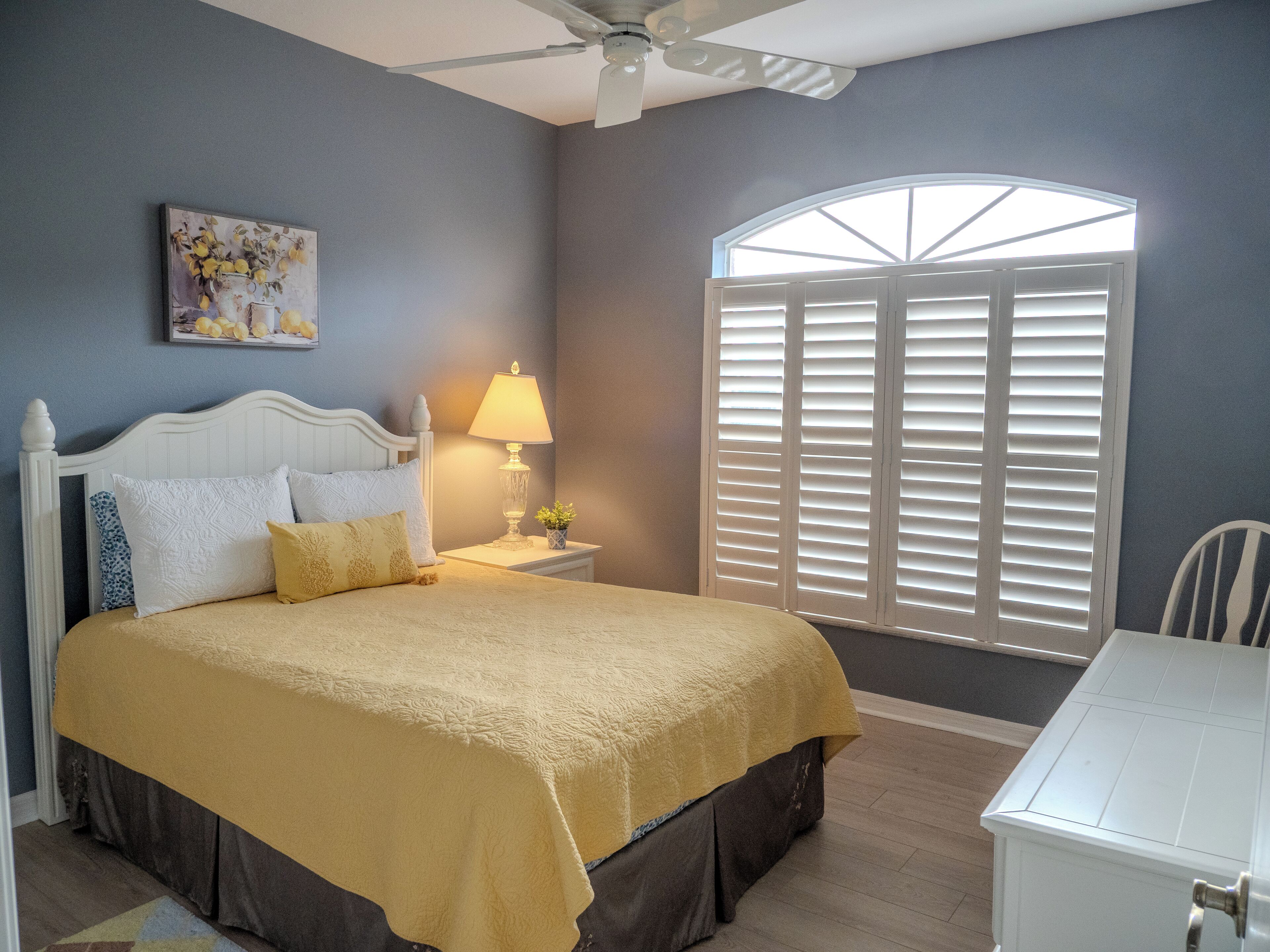 Ein Schlafzimmer mit Bett, Nachttisch, Lampe, Fenster mit Fensterläden und Deckenventilator.