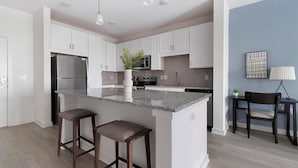 Una cocina moderna con armarios blancos, encimera de granito, refrigerador de acero inoxidable y microondas empotrado. Hay dos taburetes de bar y una planta en maceta en la isla.