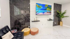 Uma sala de estar moderna com uma televisão grande presa a uma parede branca, painéis de mármore preto, um sofá, dois pufes redondos e um vaso com planta.
