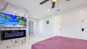 Quarto com cama, televisão fixada em parede de mármore, ventoinha no teto e portas brancas.