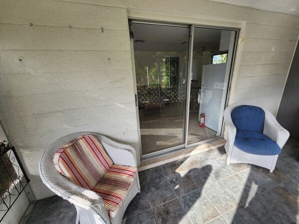 A covered patio area with two wicker chairs, one striped and one blue, and a sliding glass door leading to an outdoor seating area. - Paradise in the heart of Old Englewood Village (Englewood)
