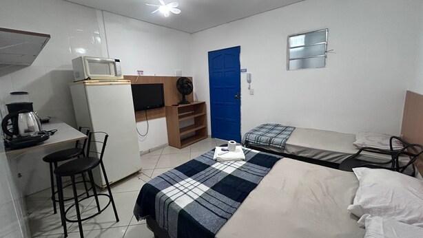 A compact hotel room with a bed, a desk with a computer, a small kitchenette, and a blue door. - Studio próximo Santana SP (São Paulo)