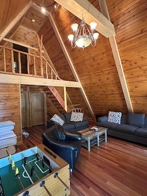 An attic room with a wooden sloped ceiling, a chandelier, a sofa set, a coffee table, and a foosball table.