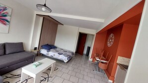 A modern living room with a sofa, a small table, a bed with a patterned bedspread, a hanging light fixture, and a view into another room with a red accent wall and a clock. - Studio ideally located (Sens)