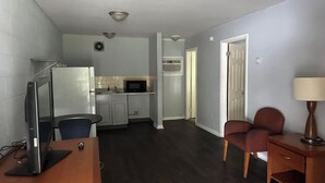 A compact living space with a kitchenette, a desk with a computer, a chair, and a lamp.