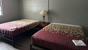 A room with two beds, each with a patterned bedspread. There is a nightstand with a lamp between the beds. The walls are painted gray, and there is a window on the left side.