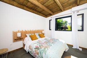 A bedroom with a wooden ceiling, a bed with a floral bedspread, a nightstand, and a window with curtains. - Villa fleur de jade (Saint-Louis)