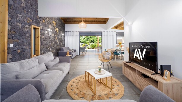 A modern living room with a stone wall, grey sofa, wooden ceiling beams, and a television displaying 'Anthony Valen 2019'. - Villa fleur de jade (Saint-Louis)