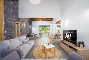 Una sala moderna con pared de piedra, sofá gris, vigas de madera en el techo y un televisor mostrando 'Anthony Valen 2019'.