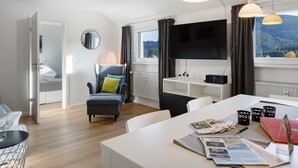 Ein modernes Zimmer mit Flachbildfernseher, einem Esstisch mit Zeitschriften, einem Sessel und Blick auf eine bewaldete Landschaft durch das Fenster.