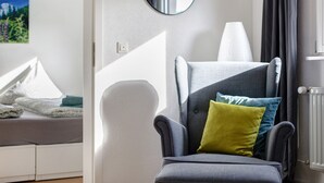 Ein modernes Zimmer mit einem grauen Sessel, einem runden Spiegel und einem Fenster mit Blick auf Grünflächen.