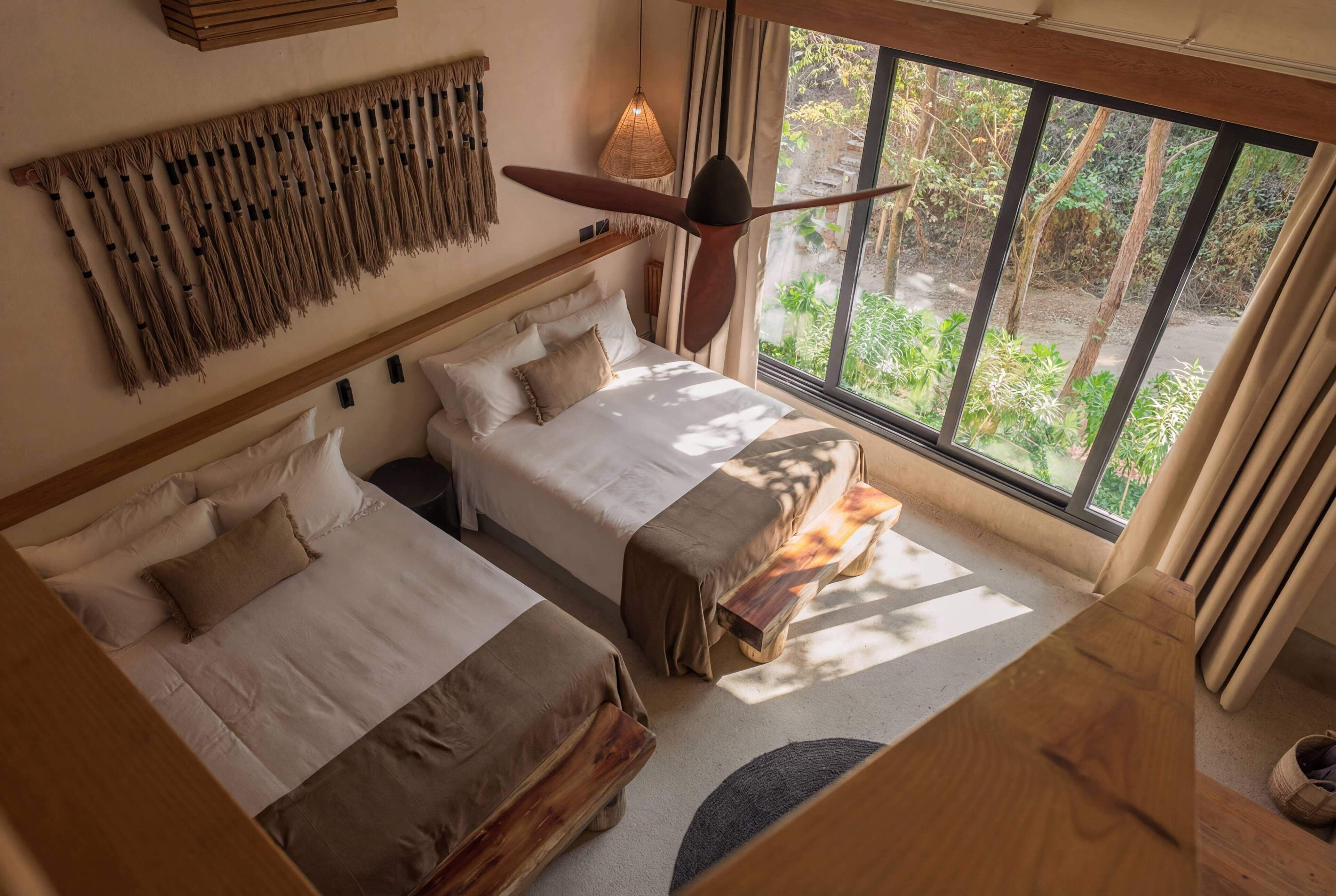 A bedroom with two beds, a ceiling fan, a large window with a view of greenery, and a woven wall hanging.