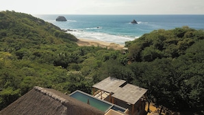 A thatched-roof building with a pool, surrounded by dense greenery and overlooking a beach with rocky outcrops.