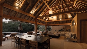 A spacious indoor area with a high ceiling and wooden beams, featuring a dining table, sofas, and a seating area with a view of the outdoors.