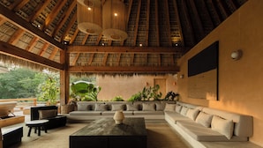 A spacious living area with a high ceiling, a large sofa set, a coffee table, and a view of greenery outside.